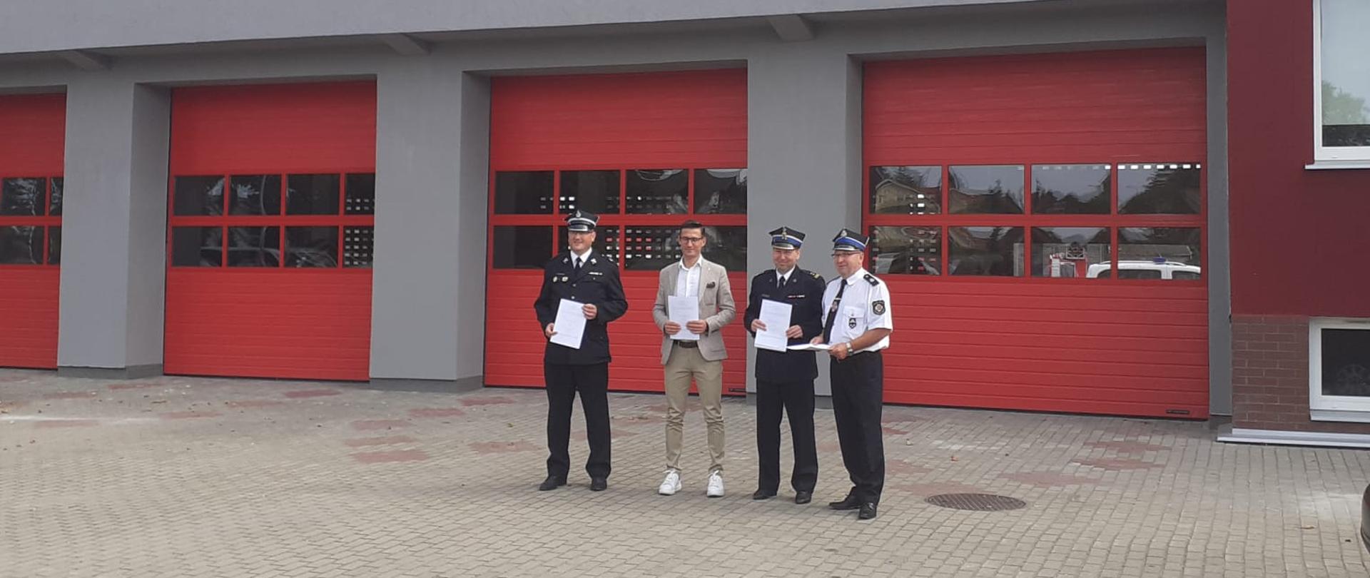 Zdjęcie przedstawia osoby stojące przed garażami straży pożarnej z podpisanym porozumieniem o włączeniu do KSRG OSP Warnice.