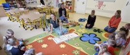 Grupa przedszkolna słuchająca lekcji prowadzonej przez pracowników powiatu ełckiego, siedzi na kolorowym dywanie wokół prowadzących 