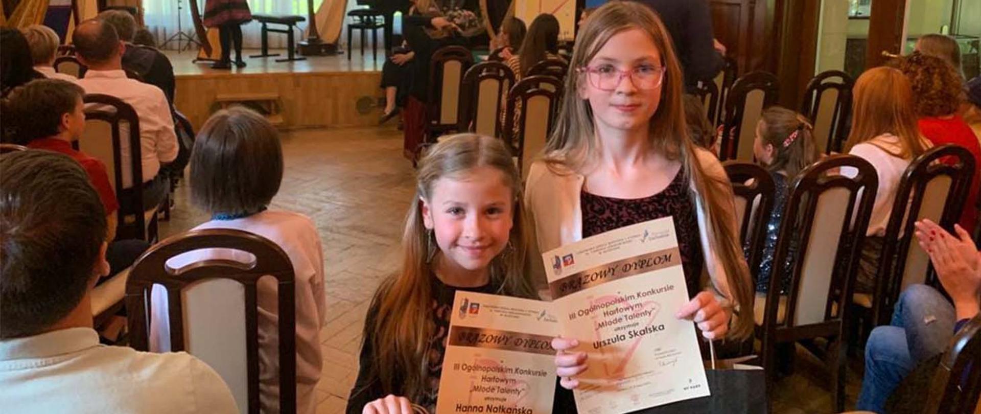 Dwie dziewczynki stoją w tyle pomieszczenia, plecami zwrócone do sceny i pozostałych osób, obie trzymają w rękach dyplomy.