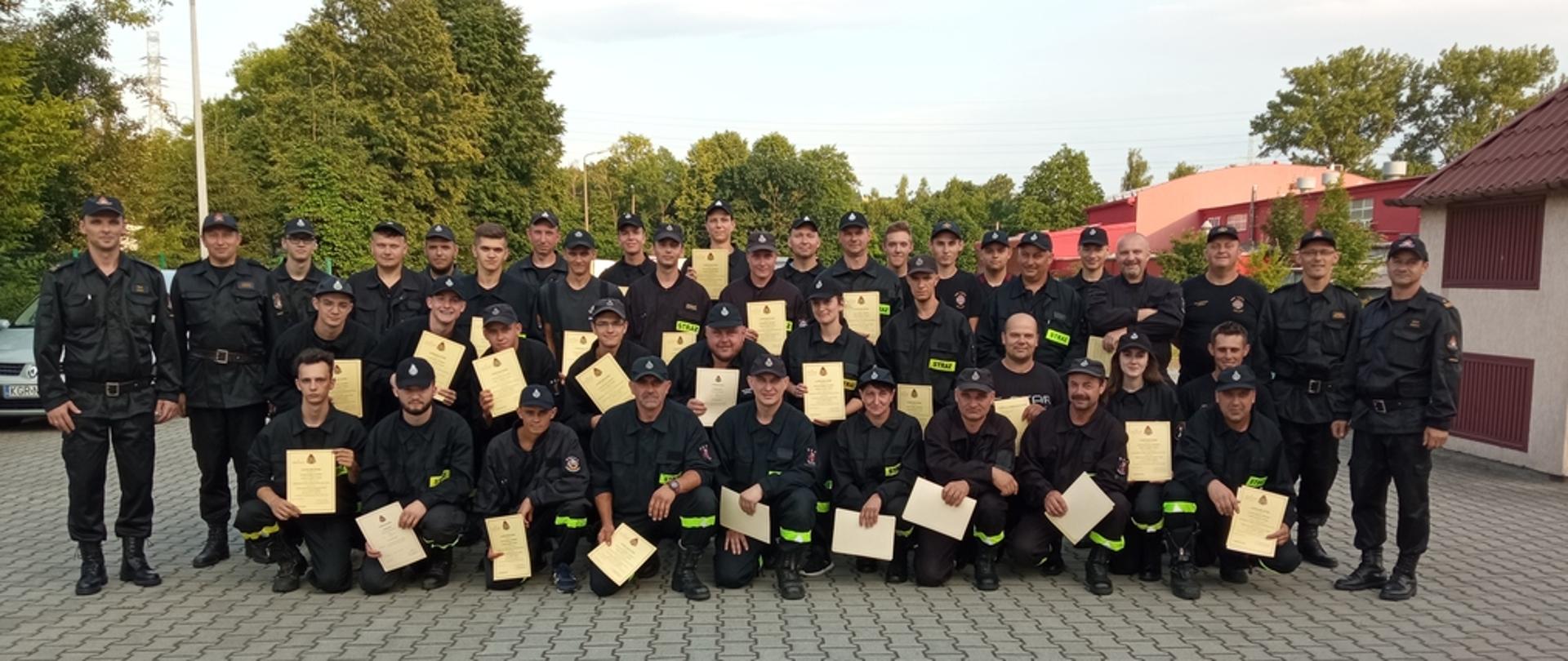 Na zdjęciu strażacy z Ochotniczych Straży Pożarnych po egzaminie kończącym szkolenie podstawowe strażaków ratowników OSP oraz komisja egzaminacyjna składająca się ze strażaków z PSP. Strażacy ubrani w ubrania koszarowe i dowódczo-sztabowe koloru czarnego w czarnych czapkach.