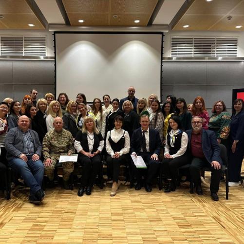 Ustawiona w trzy rzędy grupa kobiet i mężczyzn pozuje do zdjęcia na tle ściany z pustym ekranem