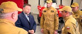 Bliskie ujęcie skupione na twarzach uczestników wizyty. Minister Marcin Kierwiński słucha wyjaśnień bryg. Daniela Myszki. Strażacy mają na sobie piaskowe mundury z naszywkami "Państwowa Straż Pożarna" oraz polskimi flagami na ramionach. W tle widoczny jest fragment czerwonej łodzi ratunkowej na przyczepie.