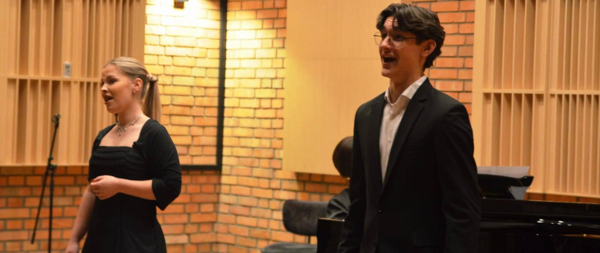 Wokalistka i wokalista w galowych strojach. W tle pianistka przy fortepianie