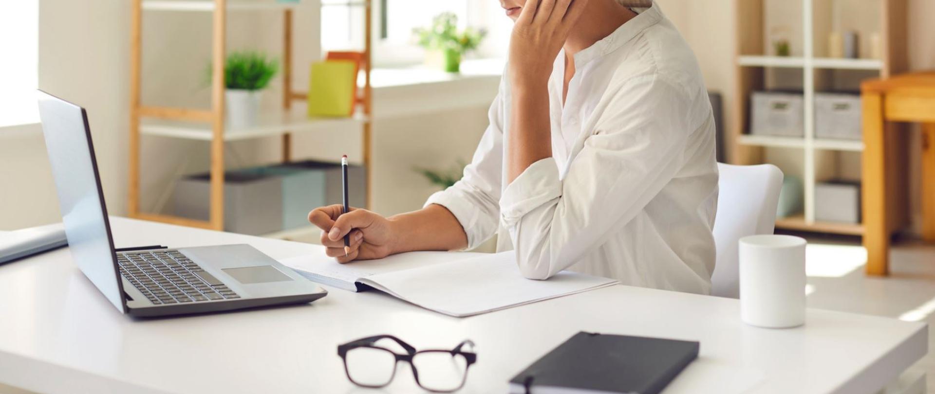 Kobieta w białej koszuli przy białym biurku, patrzy na otwarty laptop, zapisuje ołówkiem notatki w zeszycie, obok leżą okulary i biały kubek.