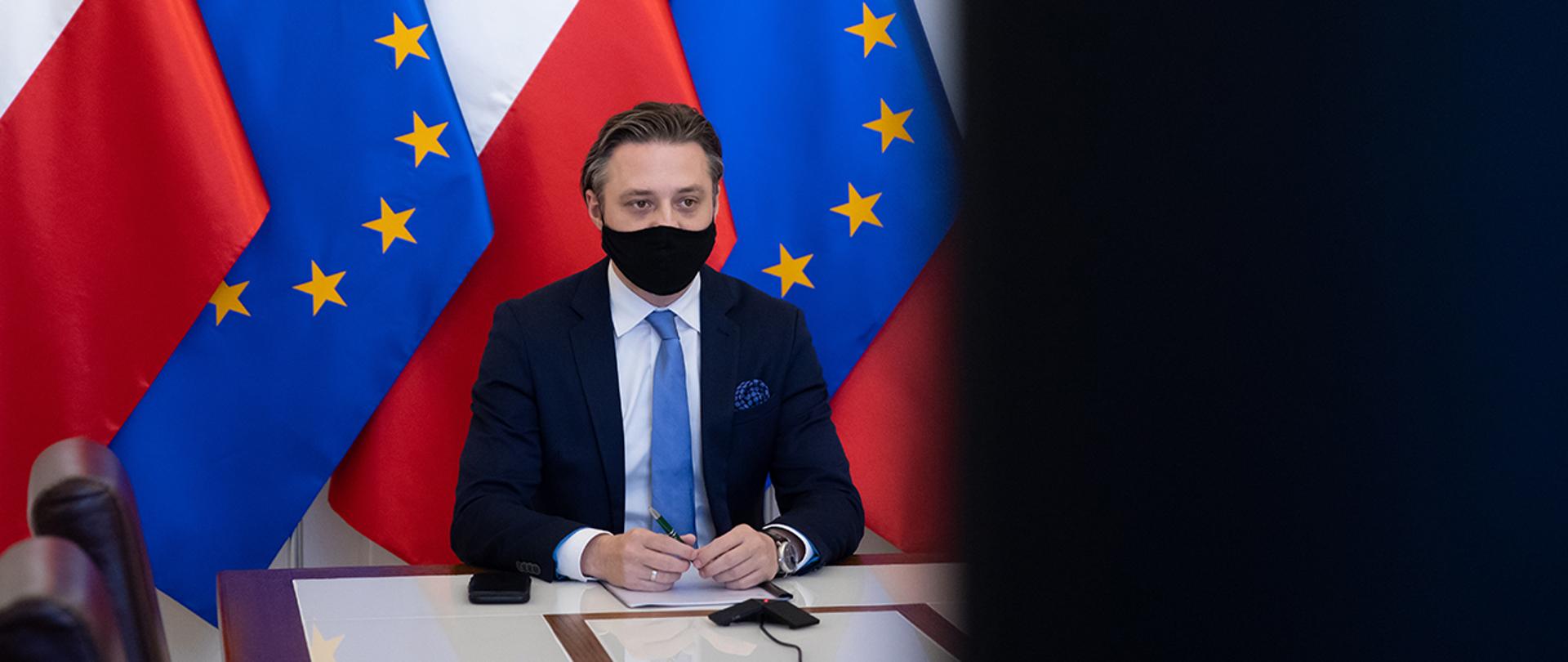 Na zdjęciu: wiceminister Bartosz Grodecki siedzący na tle flag Polski i Unii Europejskiej patrzy w ekran podczas wideokonferencji 