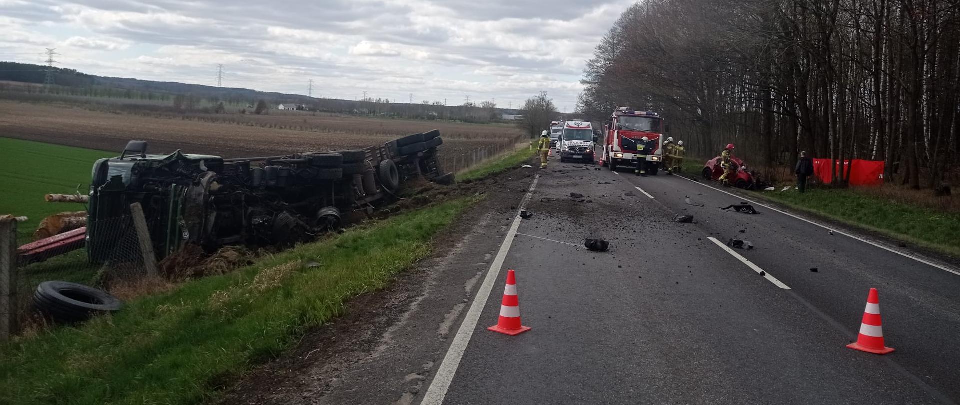 Na zdjęciu widać leżący w rowie samochód ciężarowy, samochód straży pożarnej, karetkę pogotowia oraz strażaków.