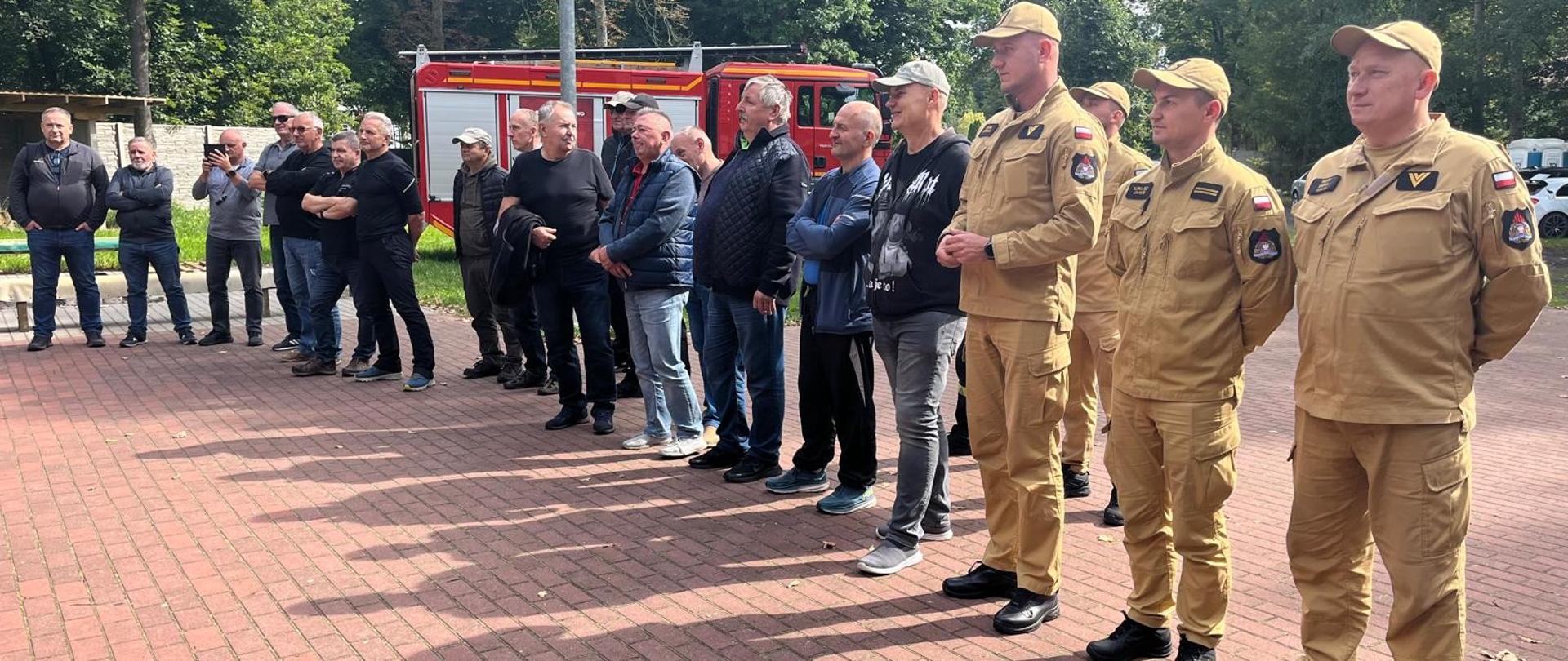 Szeroki kadr przedstawia długą linię mężczyzn stojących na placu wyłożonym czerwoną kostką brukową, rzucających długie cienie w świetle słonecznym. Na pierwszym planie po prawej stronie stoi kilku mężczyzn w jednolitych beżowych mundurach i czapkach, a w głębi widać czerwony samochód strażacki.