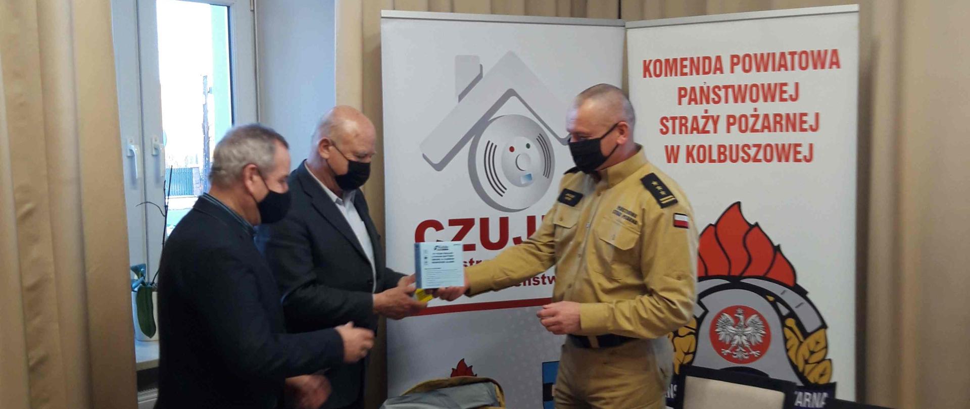 Na zdjęciu 3 mężczyzn: strażak w umundurowaniu w kolorze piaskowymi oraz dwóch w garniturach. Jednemu z mężczyzn strażak przekazuje jedną z czujek tlenku węgla. Na stole pozostałe przekazywane urządzenia oraz inne materiały. Zdjęcie w pomieszczeniu z dużym oknem i długimi zasłonami. W rogu banery z napisami: Komenda Powiatowa Państwowej Straży Pożarnej w Kolbuszowej wraz z logo PSP oraz Czujka na straży Twojego bezpieczeństwa.