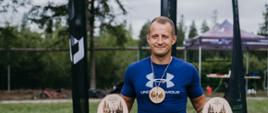 Mężczyzna uczestniczący w zawodach biegowych, trzymający w dłoniach zdobyte trofea. Mężczyzna soi na trawie, a za nim na drugim planie widoczne jest podium.