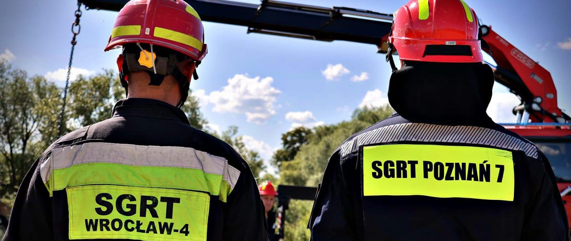 Dwóch strażaków w hełmach i ubraniach ochronnych stoi tyłem do obiektywu. Na ich ubraniach widoczne są oznaczenia „SGRT WROCŁAW-4” oraz „SGRT POZNAŃ 7”. W tle znajduje się samochód ratowniczy z wysięgnikiem, a całość jest przedstawiona w plenerze przy słonecznej pogodzie.