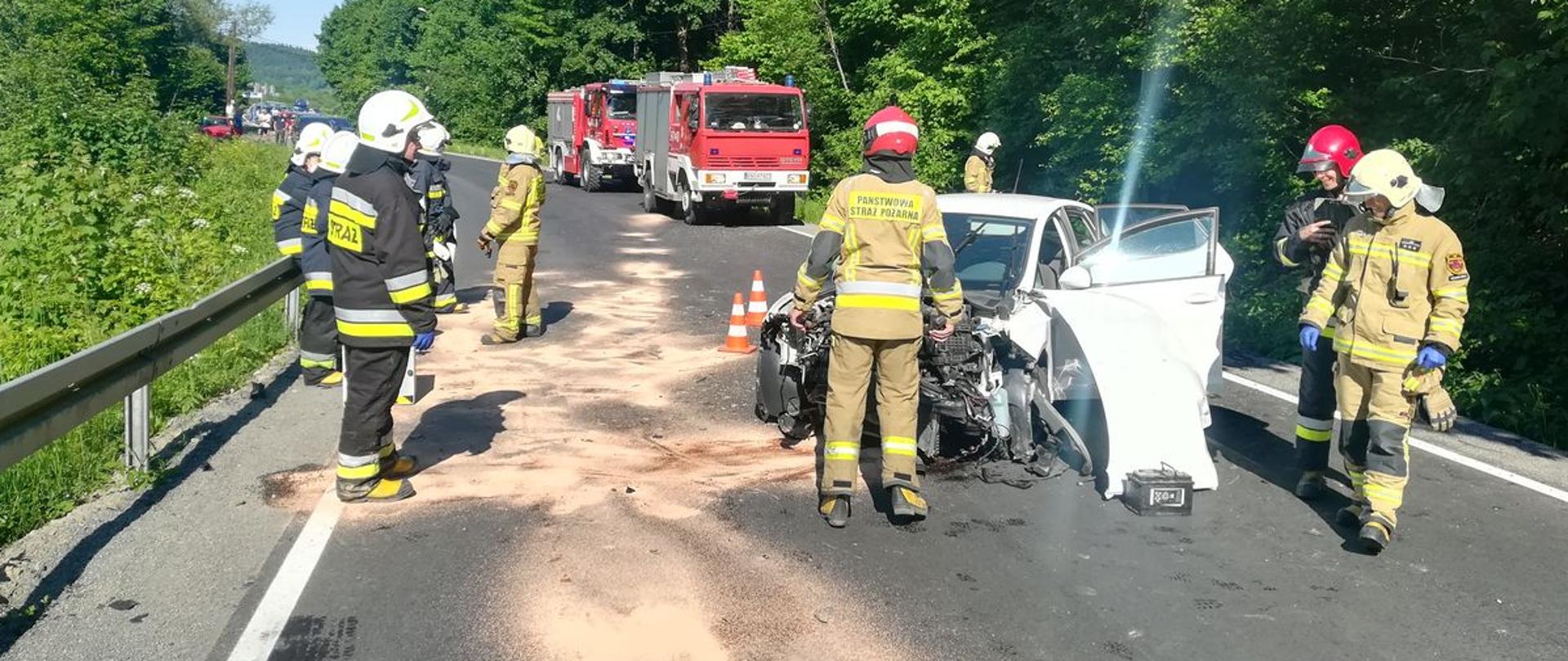 Samochód osobowy białego koloru z rozbitym przodem stoi w poprzek drogi. Wokół niego stoją strażacy. Na drodze zasypane sorbentem płyny eksploatacyjne. W tle samochody pożarnicze.