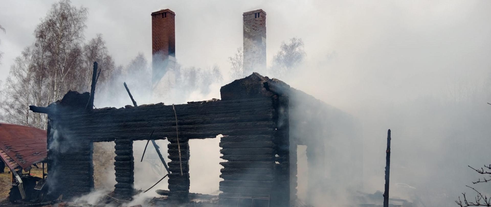 Widok na spalony drewniany budynek mieszkalny. Wokół niego kłęby dymu.