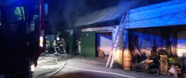 Zdjęcie przedstawia strażaków gaszących pożar garażu. W tle widać budynki gospodarcze, linie gaśnicze oraz samochód.