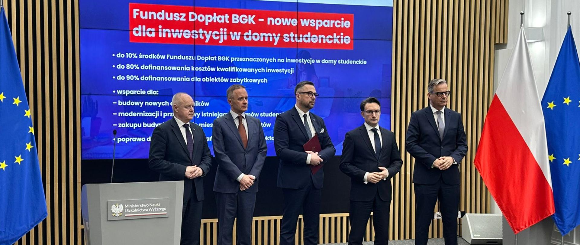 W dużej sali stoi pięciu mężczyzn w garniturach pośrodku minister Kulasek, za nimi ściana z cienkich pionowych listewek z wbudowanym wielkim ekranem z napisem Fundusz Dopłat BGK - nowe wsparcie dla inwestycji w domy studenckie, po bokach flagi Polski i UE.