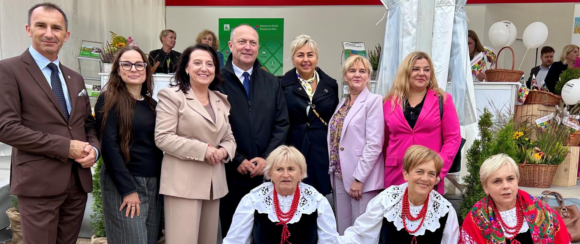 Pięć kobiet i dwóch mężczyzn pozuje do zdjęcia na tle stoiska informacyjnego KRUS pod białym namiotem.. W pierwszym rzędzie przysiadły trzy kobiety w strojach ludowych.