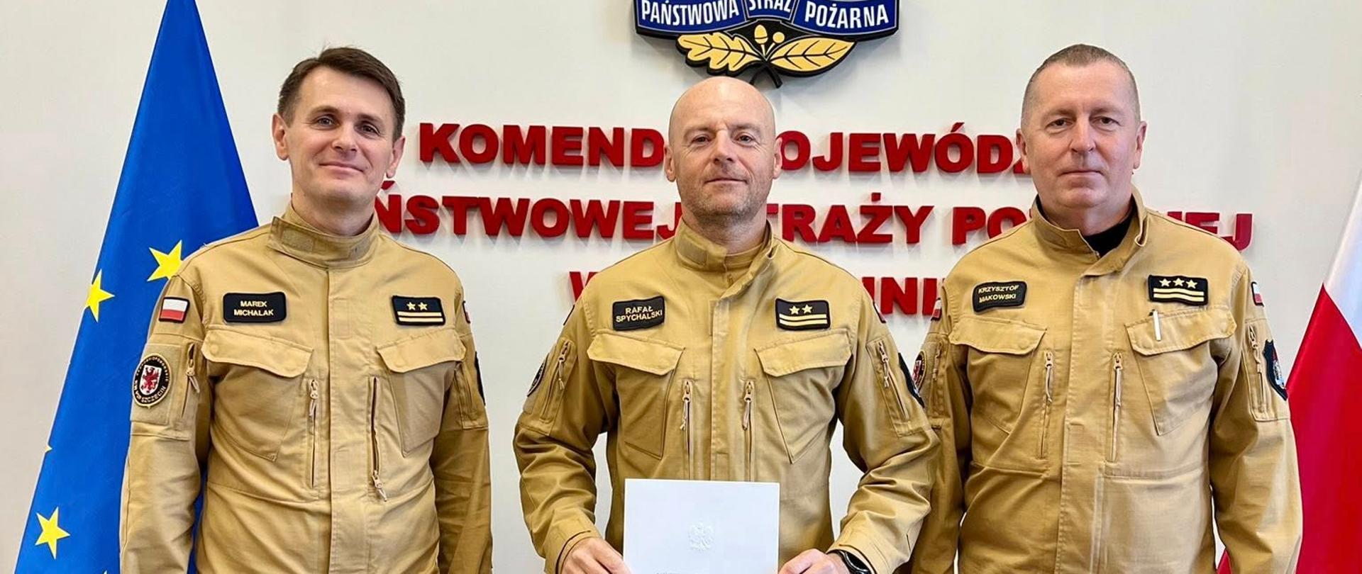 Na zdjęciu stoją trzej mężczyźni w piaskowych mundurach Państwowej Straży Pożarnej. Wszyscy mają czarne buty i naszywki na ramionach. Środkowy mężczyzna trzyma w dłoniach biały dokument z napisem „Państwowa Straż Pożarna”. W tle widnieje ściana z godłem Państwowej Straży Pożarnej – orłem w czerwonym płomieniu, otoczonym złotym wieńcem – oraz napis w czerwonych literach: „KOMENDA WOJEWÓDZKA PAŃSTWOWEJ STRAŻY POŻARNEJ”. Po lewej stronie stoi flaga Unii Europejskiej, a po prawej flaga Polski. Zdjęcie zostało wykonane w pomieszczeniu urzędowym, w jasnym świetle