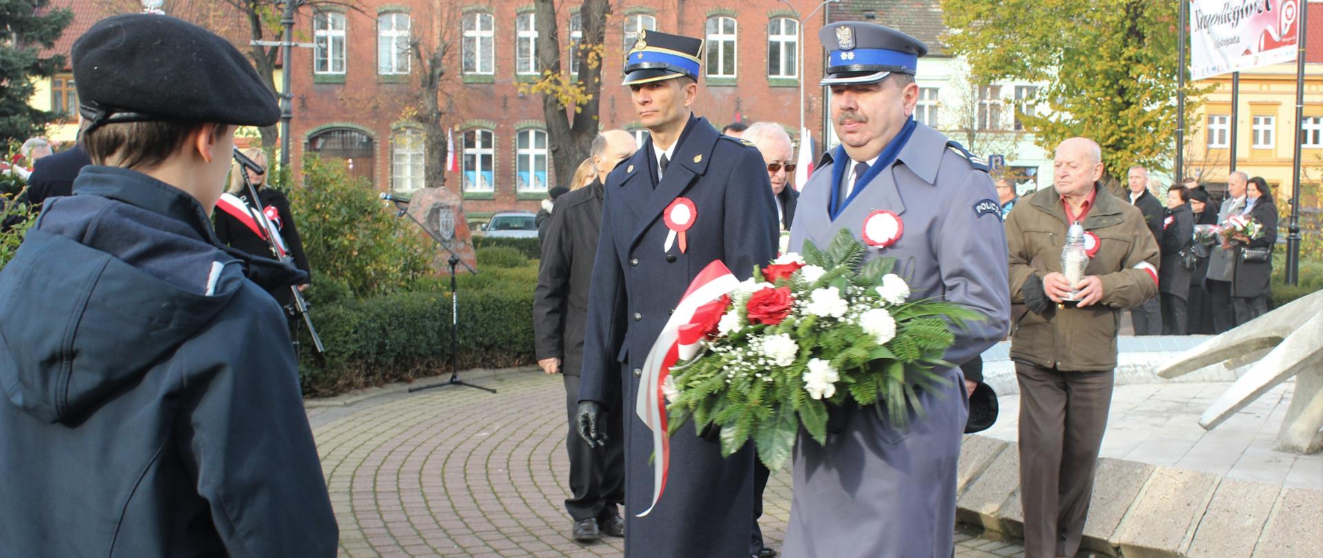 Panowie Komendanci Straży Pożarnej i Policji w płaszczach wyjściowych z wieńcem.
