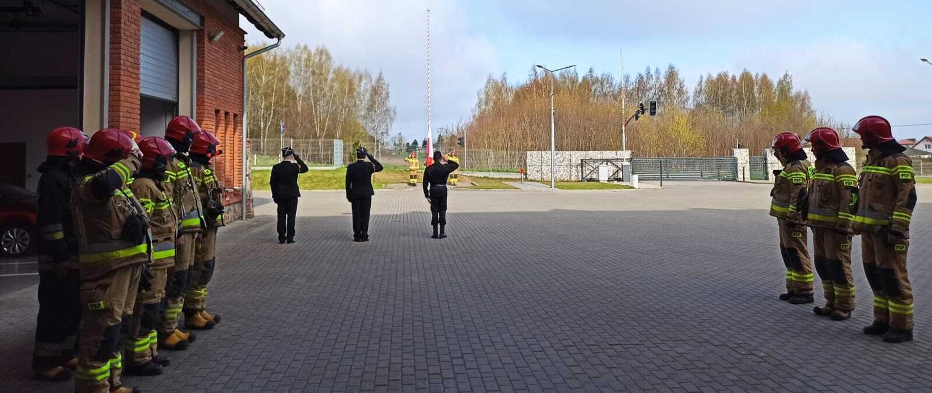 Zdjęcie przedstawia strażaków Komendy Powiatowej Państwowej Straży Pożarnej w Gołdapi podczas podnoszenia flagi Rzeczypospolitej Polskiej przy akompaniamencie hymnu państwowego z okazji Dnia Flagi Państwowej Rzeczypospolitej Polskiej. Dowódcy zmian, funkcjonariusze nie będący w pododdziale oraz dowódca pocztu flagowego i asystujący oddają honory przez salutowanie