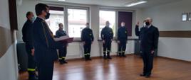 Na świetlicy komendy odbywa się uroczysta zbiórka z okazji ślubowania nowego strażaka. W tle ustawieni strażacy zmiany służbowej w ubraniach koszarowych w kolorze czarnym. Po lewej stronie stoi Komendant Powiatowy, w mundurze galowym, przyjmujący rotę ślubowania. Po prawej składający ślubowanie strażak w umundurowaniu służbowym z uniesioną do ślubowania prawą ręką. 