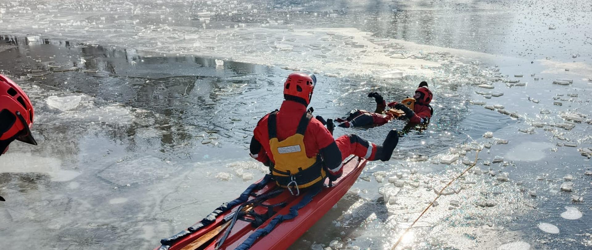 Strażak siedzący na desce lodowej podejmuje drugiego strażaka pełniącego rolę pozoranta z wody na zamarzniętym akwenie. Strażacy zabezpieczeni w środki ochrony indywidualnej i asekurowani za pomocą lin i kamizelek.
