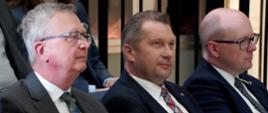 Minister Czarnek i dwóch mężczyzn w garniturach siedzą na krzesłach, za nimi inni ludzie.
