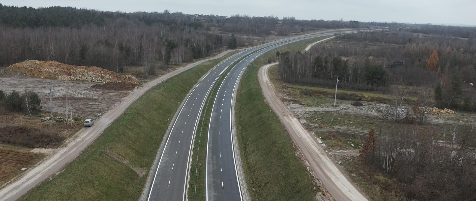 DK42 obwodnica Wąchocka - zdjęcie dwujezdniowej trasy w wykopie wykonane z drona, Droga biegnie między drzewami, zakręca w oddali w prawo. Wzdłuż drogi głównej jej obu stronach biegną wzdłuż jezdni wąskie drogi dojazdowe na nasypach. 