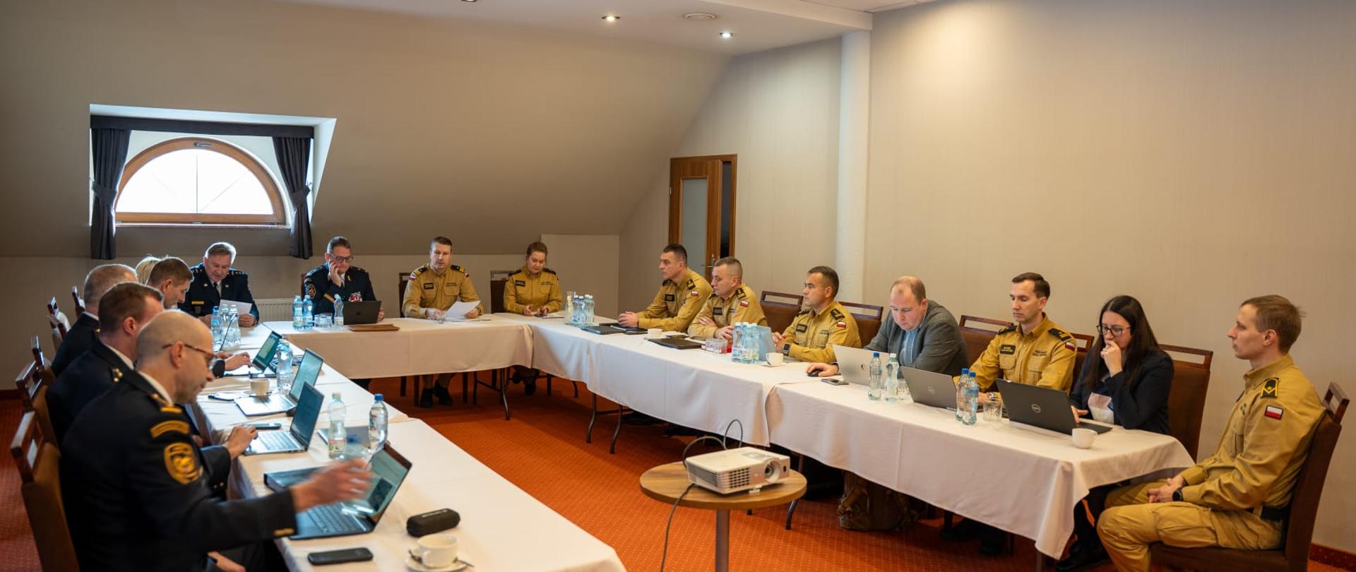 Szeroki kadr sali konferencyjnej, w której polscy strażacy oraz przedstawiciele administracji z obu państw siedzą wokół stołu w kształcie podkowy, pracując na laptopach i analizując materiały podczas spotkania polsko-czeskiej grupy roboczej.