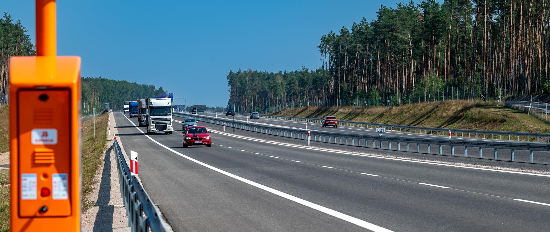 Zdjęcie przedstawia autostradę A2 biegnącą przez zalesiony teren. Po lewej stronie widoczny jest pomarańczowa kolumna alarmowa, która znajduje się na poboczu drogi. Na autostradzie poruszają się różne pojazdy, w tym ciężarówki i samochody osobowe, w obu kierunkach. Droga jest szeroka, z wyraźnymi pasami ruchu oraz barierkami ochronnymi z każdej strony. Na horyzoncie widać las i niebieskie niebo.
