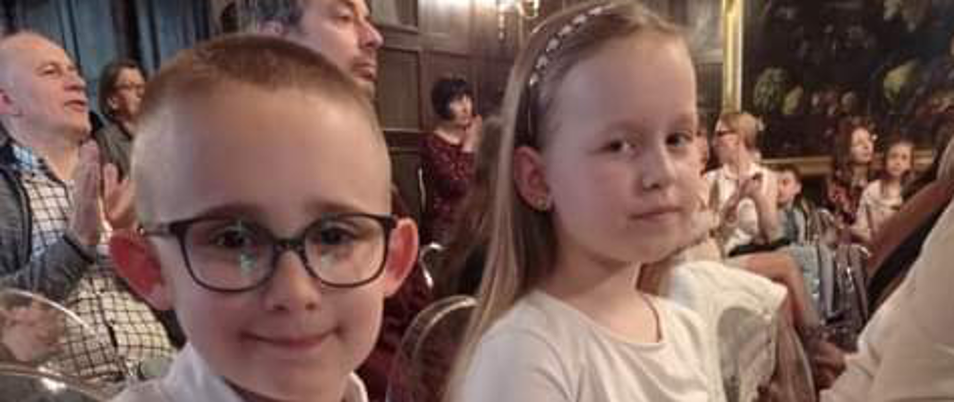 Dziewczynka i chłopiec w okularach ubrani na biało.Za nimi inni ludzie