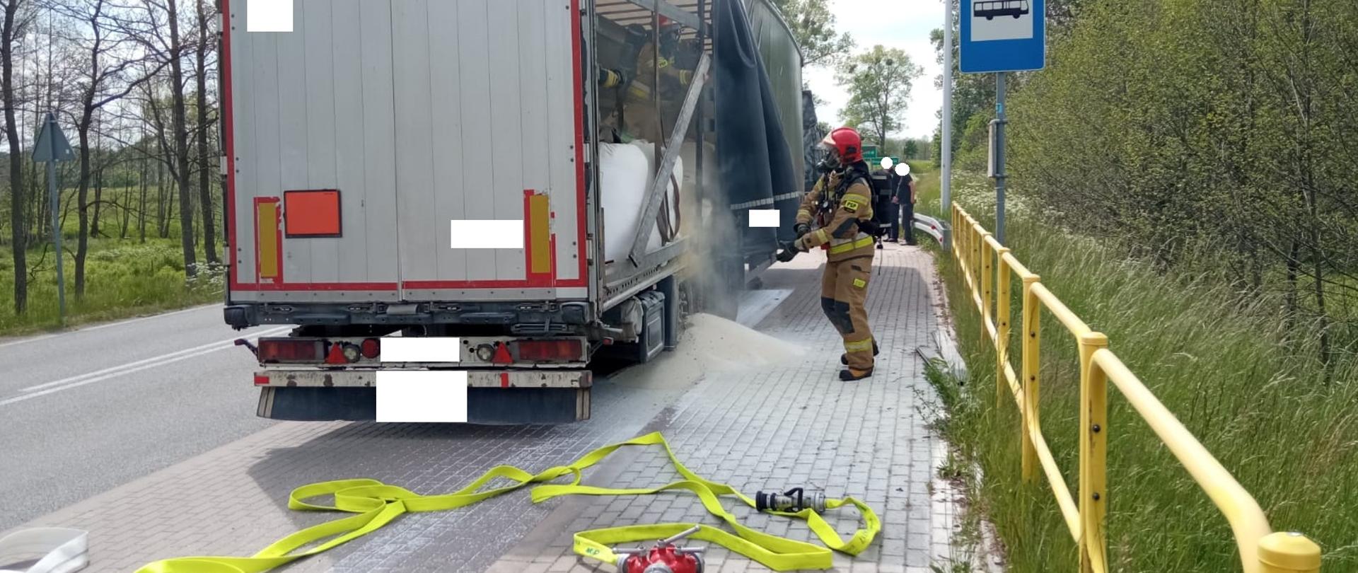 Zdjęcie przedstawia naczepę z nawozami umieszczonymi w wielkogabarytowych workach, gdzie dwa z nich były objęte ogniem. Obraz przedstawia jednego strażaka stojącego poza pojazdem oraz dwóch strażaków będących w naczepie, którzy są odpowiednio zabezpieczeni m.in. w sprzęt ochrony układu oddechowego. Na dole fotografii widać również rozwiniętą linię gaśniczą.
