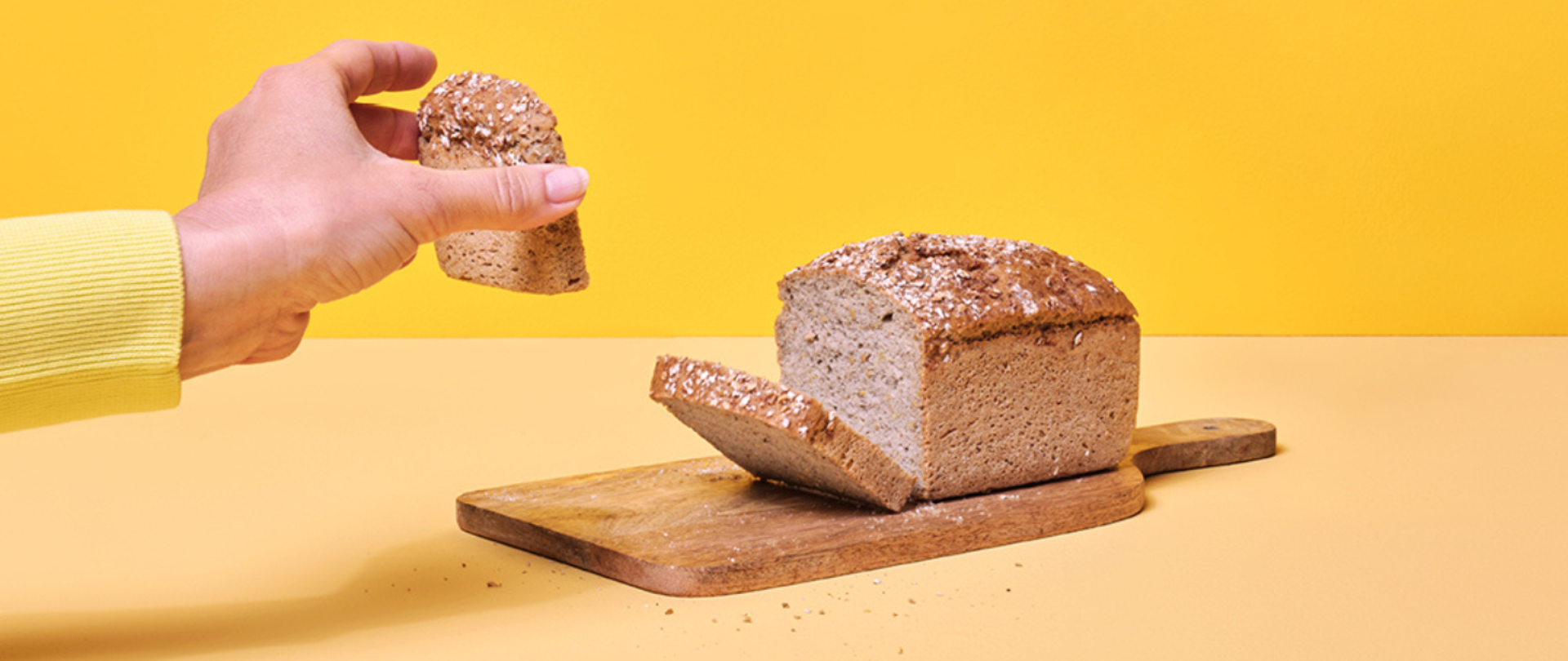Na fotografii widoczna dłoń trzymająca kromkę chleba, na środku widoczny bochenek chleba na drewnianej desce do krojenia, tło w kolorze żółtym. 
