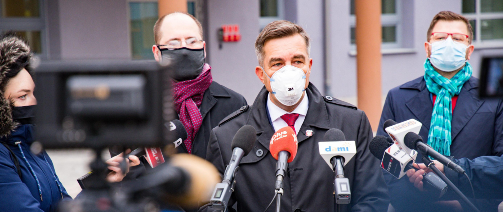 Briefing prasowy. Wiceminister Zbigniew Gryglas stoi przy mikrofonach. Za nim stoi dwóch mężczyzn, z boku po lewej kobieta w kapturze - wszyscy noszą maseczki.