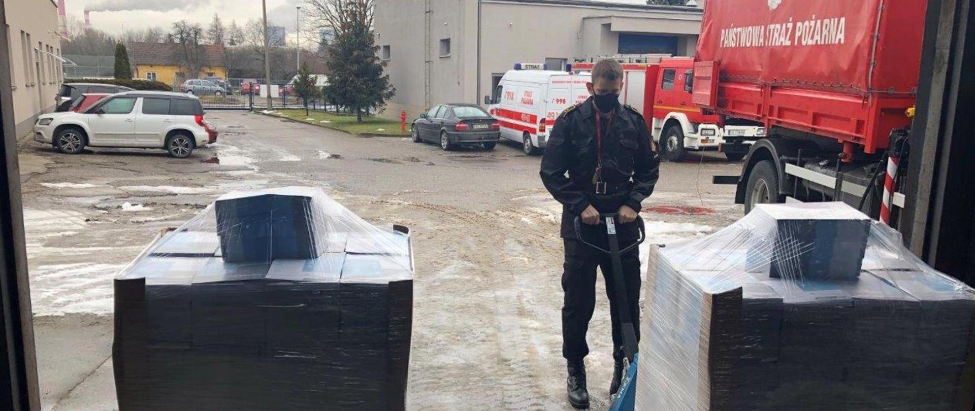 Parking między budynkami. Samochody Państwowej Straży Pożarnej. Dwie palety ulotek obok strażak.