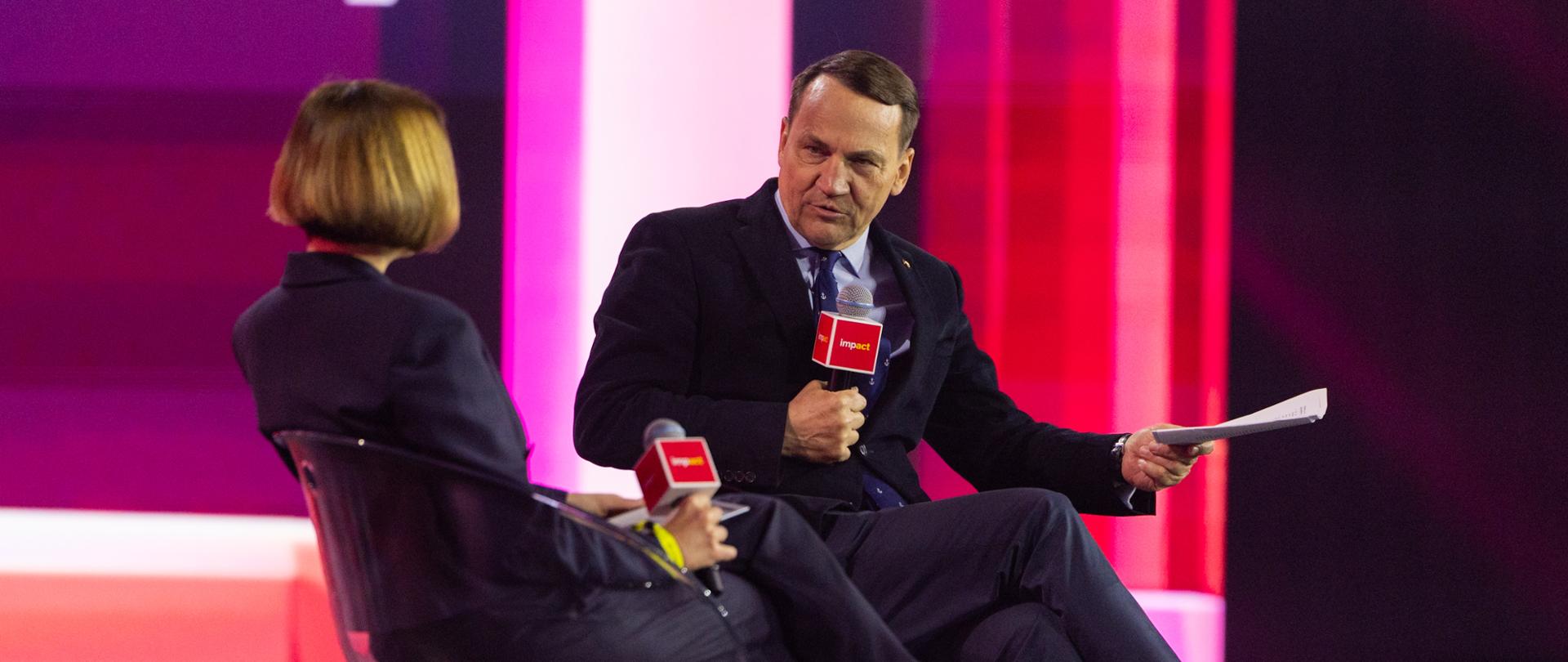Back of a woman interviewing Deputy Prime Minister Sikorski, seated on stage during a panel discussion. Politician, wearing a dark suit and blue tie, holds a microphone and documents, while the illuminated “impact” sign in pink and red colors is visible in the background.