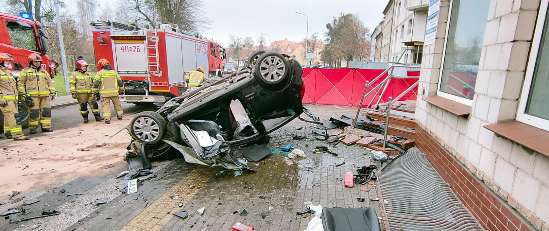 Rozbite auto przewrócone na dach. Porozrzucane elementy rozbitego samochodu. Z lewej strony na drodze ustawione samochody strażackie i strażacy.