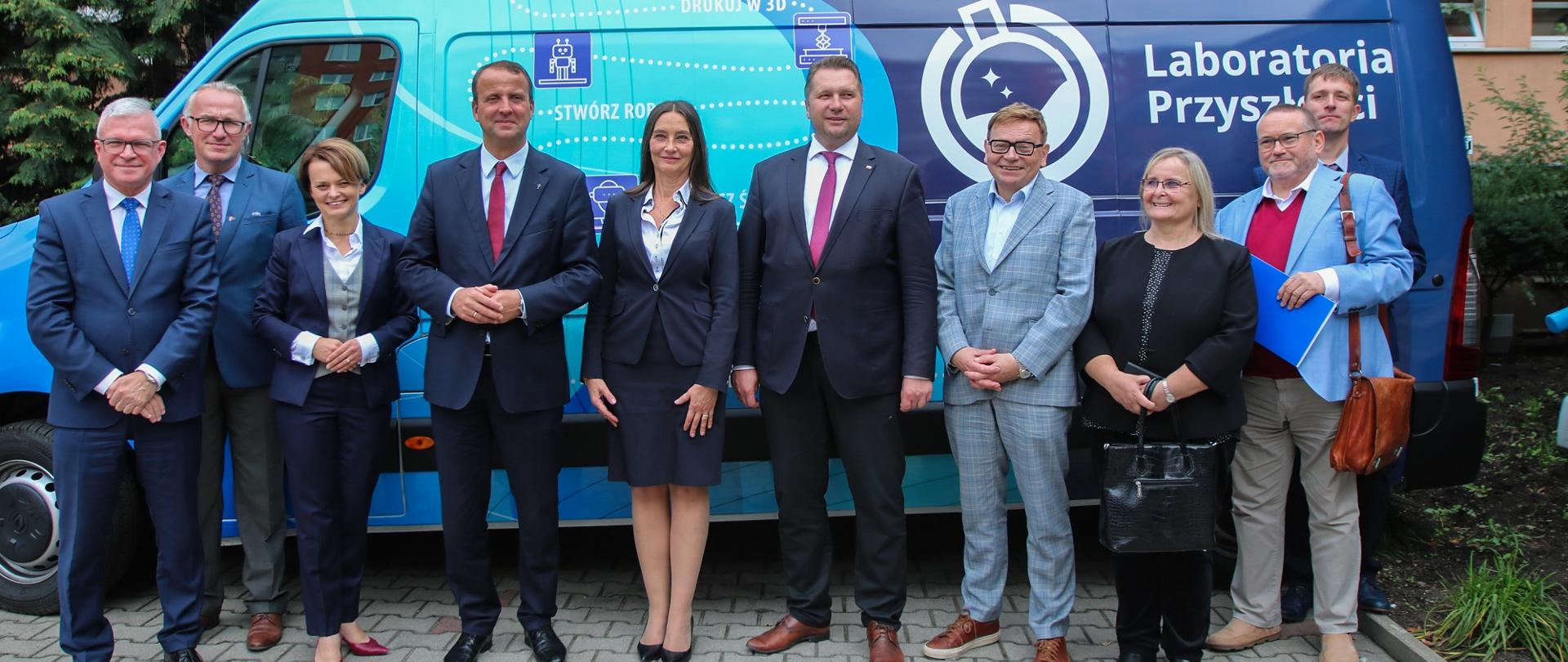 Minister w otoczeniu grupy elegancko ubranych ludzi stoi przed szkoła, za nimi niebieska furgonetka z napisem Laboratoria Przyszłości.