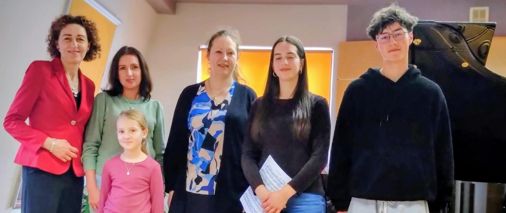 Na zdjęciu widać sześć osób stojących obok siebie w sali z fortepianem. Prawdopodobnie jest to sala koncertowa lub prób. W rzędzie stoją: Kobieta po lewej, ubrana w czerwoną marynarkę. Kobieta w zielonym swetrze, obok której stoi Dziewczynka w różowej bluzce z długim rękawem. Kobieta pośrodku w czarnej spódnicy i czarnej bluzce z niebieskim wzorem. Starsza dziewczyna w czarnym swetrze i jasnych dżinsach, trzymająca w rękach złożoną kartkę nut. Chłopak po prawej stronie, w czarnej bluzie z kapturem i szarych spodniach dresowych. Ma ciemne, kręcone włosy. W tle, po prawej stronie, widoczny jest czarny fortepian koncertowy. Sala ma jasne ściany i okna, przez które wpada dzienne światło.