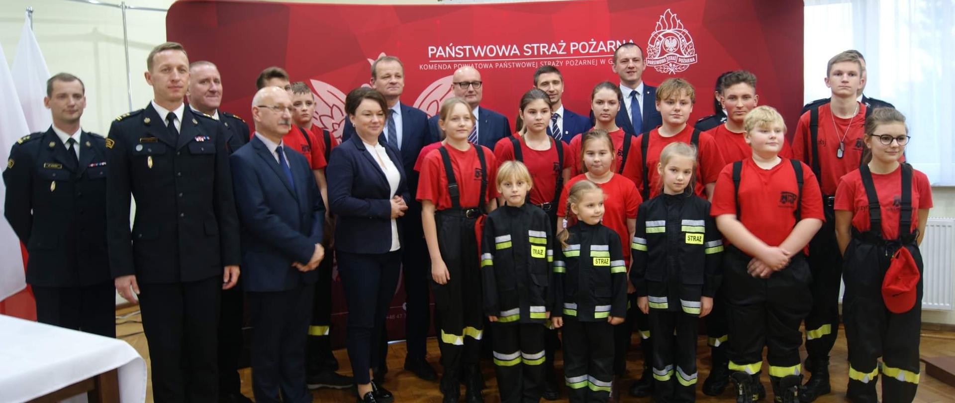 Wspólne zdjęcie dzieci, strażaków i osób wręczających promesy na tle czerwonej ścianki.