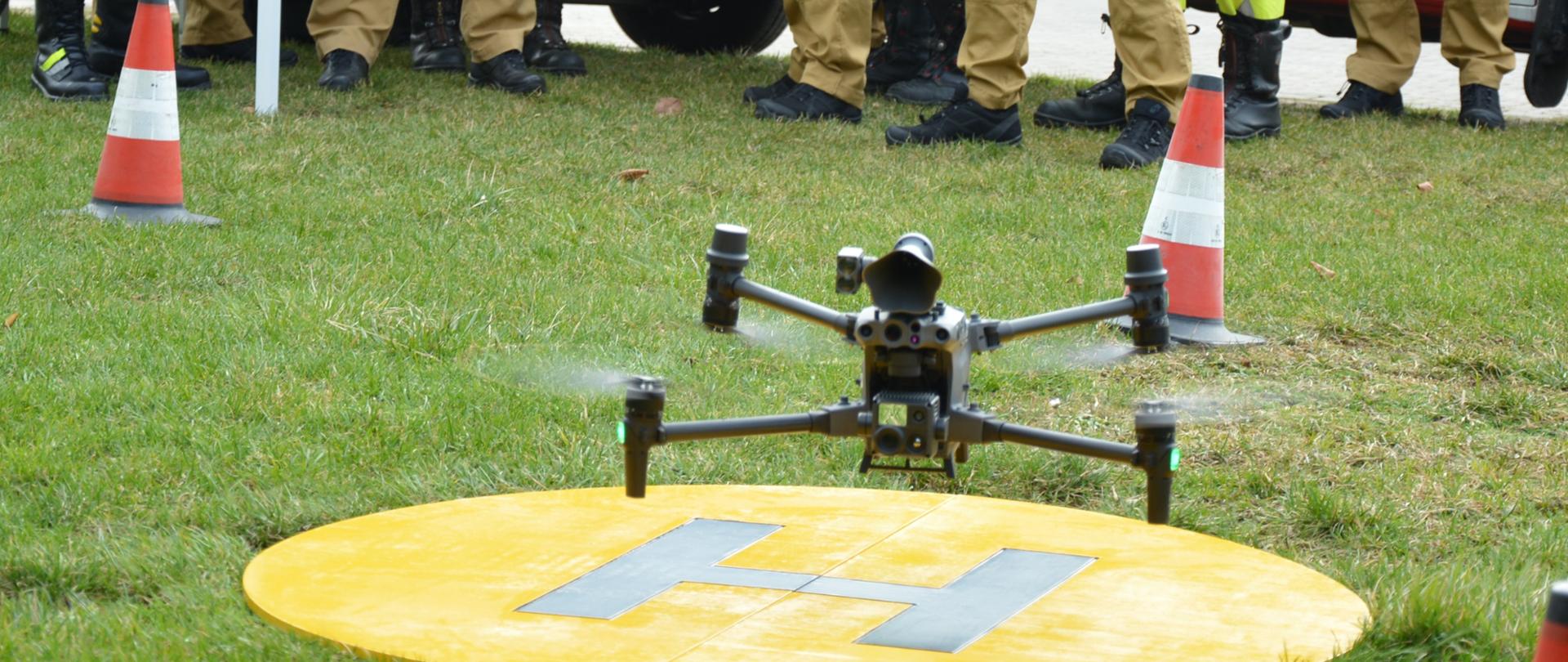 Dron startujący z miejsca wyznaczonego na lądowisko. Uczestnicy szkolenia przyglądają się dronowi.