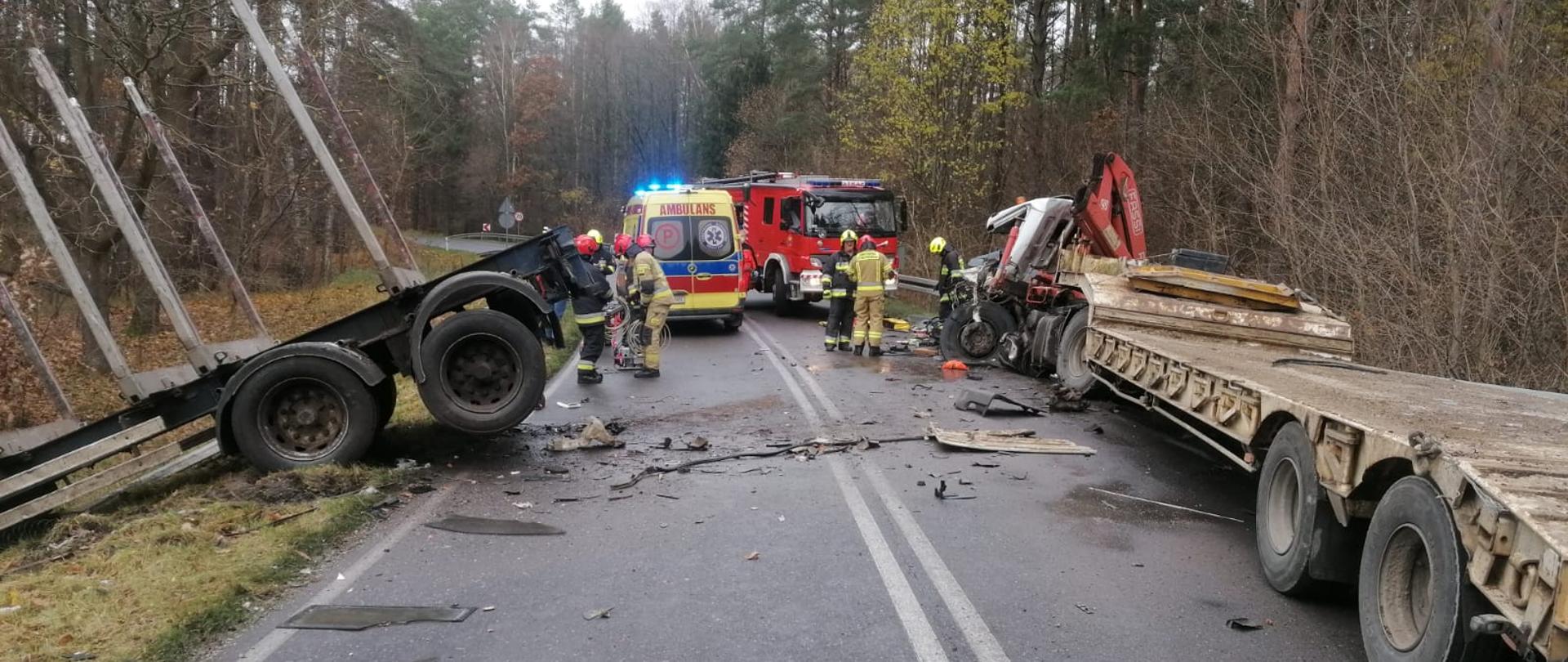 Zdjęcie przedstawia drogę dwupasmową wraz z barierkami ochronnymi na których zatrzymał się samochód ciężarowy z naczepą. Samochód koloru białego jest doszczętnie rozbity. Przed i dookoła autem znajdują się elementy z rozbitego pojazdu. Przed pojazdem znajduje się karetka pogotowia, wóz strażacki oraz strażacy. Po lewej stronie znajduję się rozbita naczepa z drugiego pojazdu.