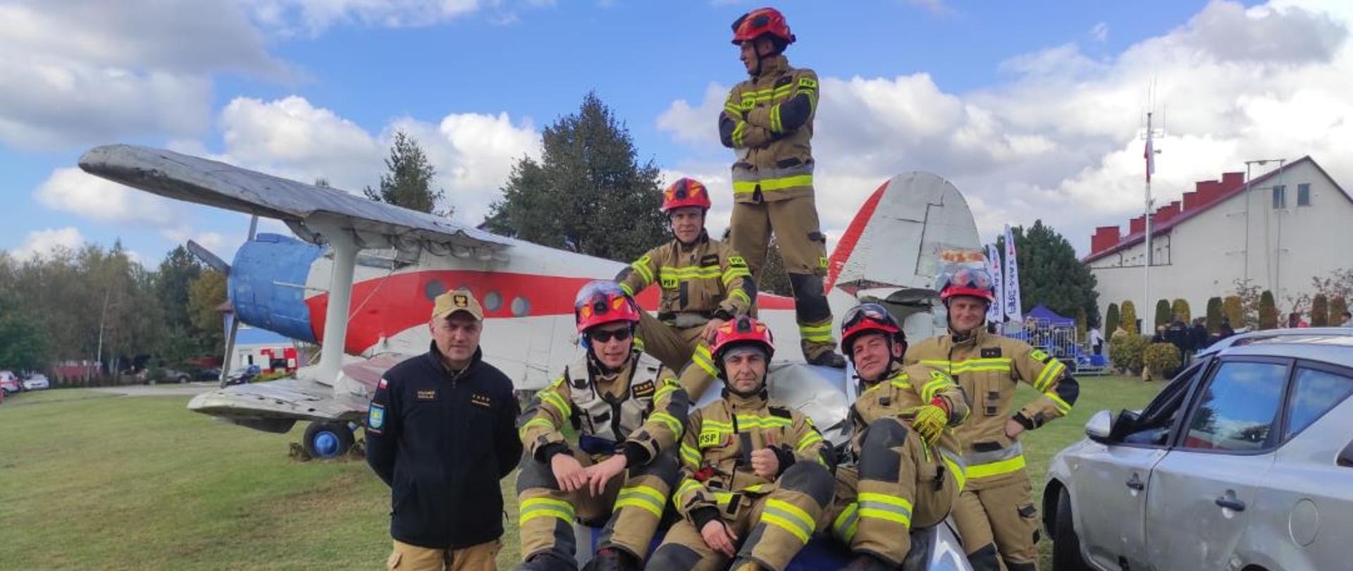 Kolorowa fotografia wykonana w słoneczny dzień na zewnątrz. Przedstawia strażaków w ubraniach specjalnych i hełmach oraz jeden w ubraniu służbowym znajdujących się na wraku samochodu osobowego po zawodach w ratownictwie technicznym. Jeden z nich posiada okulary i kamizelkę kierującego działaniem ratowniczym. W tle wrak samolotu. 