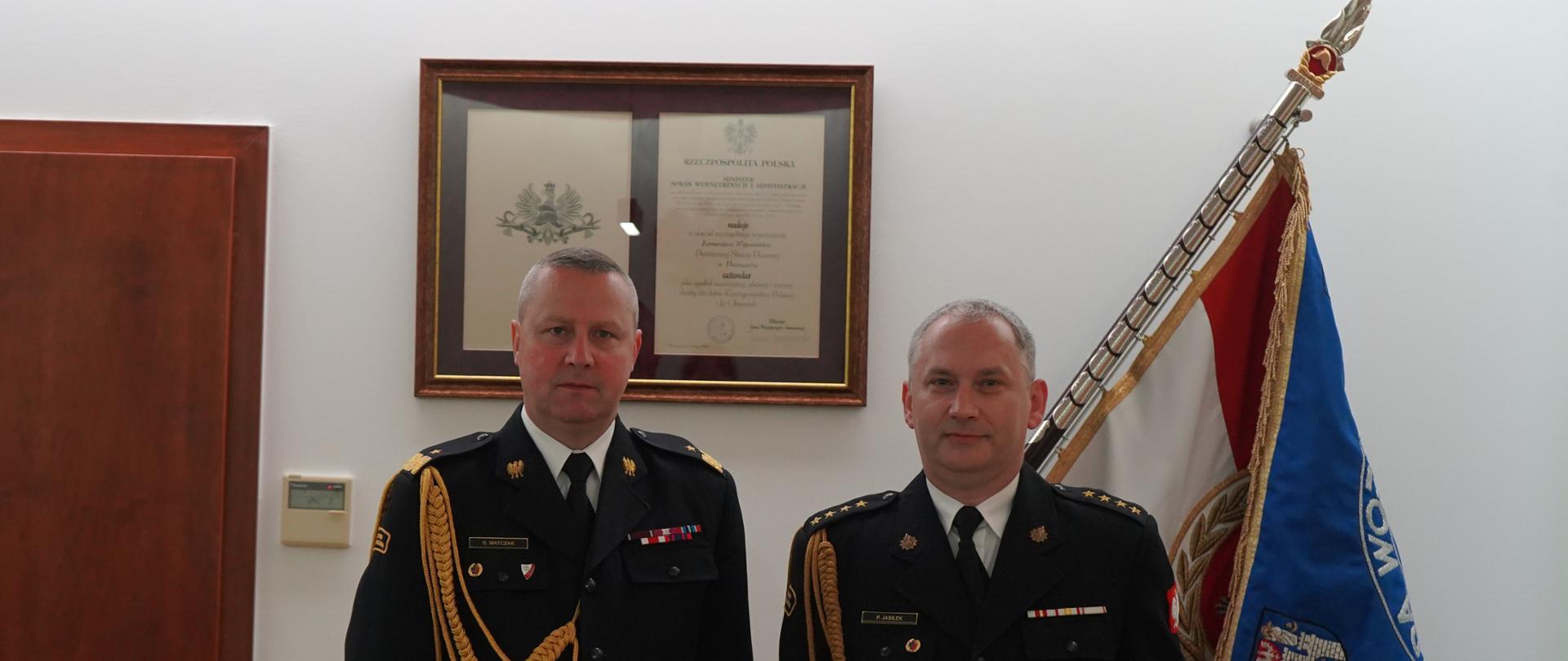 zdjęcie dwóch strażaków, jeden trzyma czerwoną teczkę z orłem, w tle sztandar