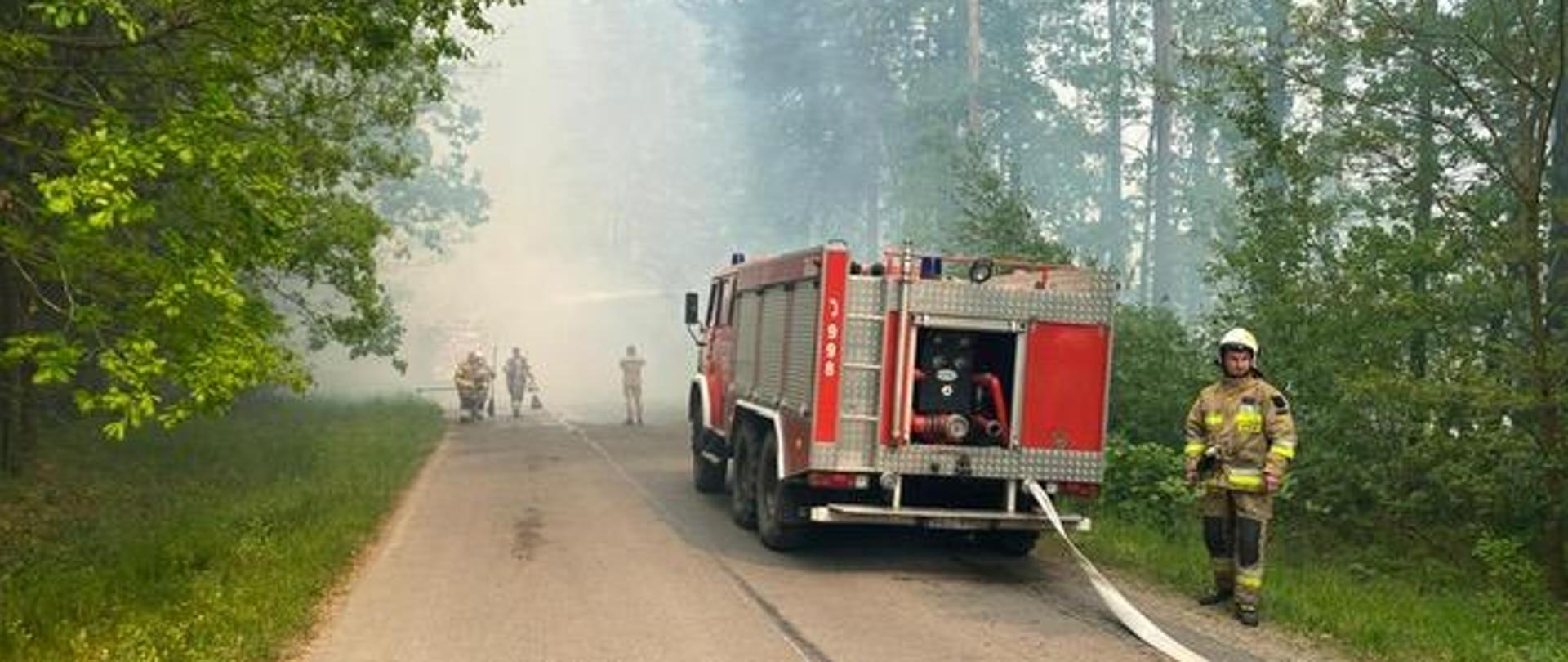 Zdjęcie przedstawia drogę w lesie i stojący na niej samochód pożarniczy. W głębi silnie zadymiony las i ratownicy ze sprzętem gaśniczym.