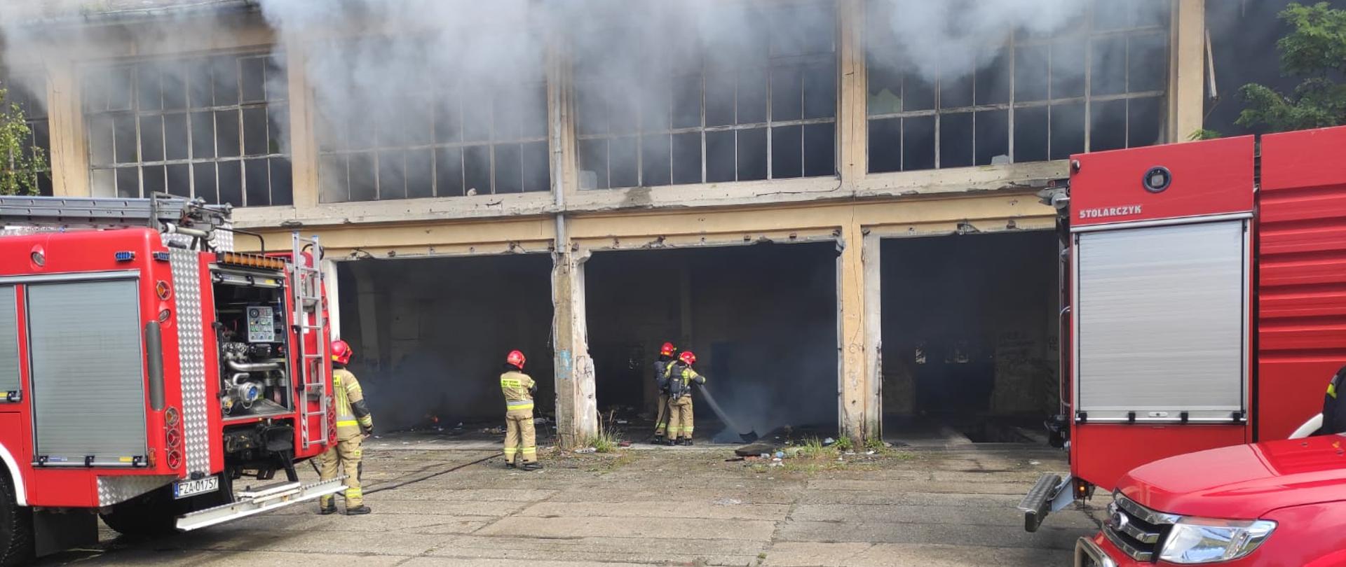 Strażacy gaszą pożar śmieci w byłym budynku jednostki wojskowej. Przed budynkiem ustawione są trzy samochody strażackie. 