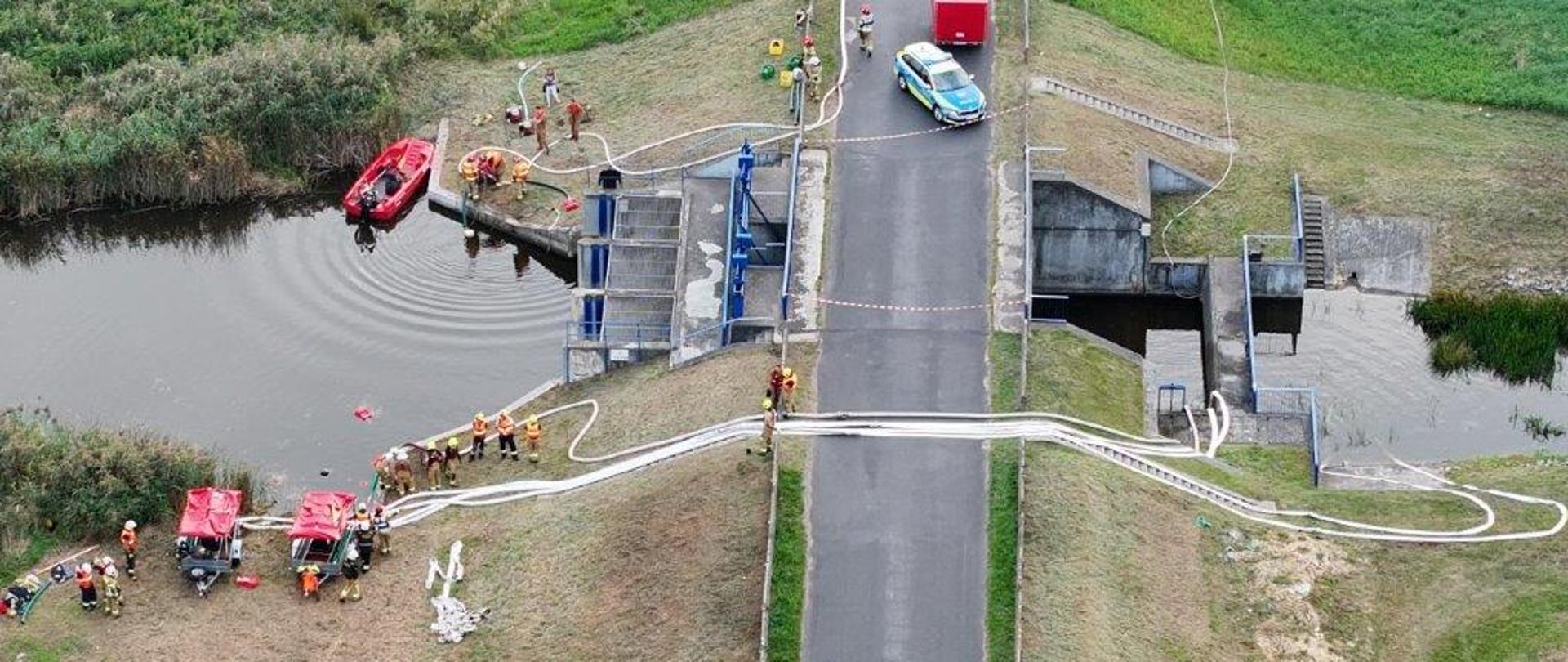 Zdjęcie przedstawia widok z lotu ptaka na akcję ratunkową lub ćwiczenia służb mundurowych, prowadzone na drodze przebiegającej przez groblę lub most nad kanałem wodnym.Na zablokowanej drodze asfaltowej widoczny jest zaparkowany radiowóz policyjny oraz biały i czerwony pojazd techniczny. Po lewej stronie akwenu, przy betonowym przepuście wodnym, znajduje się czerwona łódź motorowa oraz grupa strażaków w pomarańczowo-brązowych strojach bojowych. Druga grupa strażaków oraz rozstawiony sprzęt (w tym pompy i czerwone pojazdy ratownicze) znajdują się na trawiastym poboczu poniżej drogi.Przez jezdnię przeciągnięte są długie, białe węże strażackie, które łączą obie strony kanału rozdzielonego nasypem. Okolica jest pełna zieleni – wokół rozciągają się łąki, pola oraz gęste trzcinowiska przy brzegu wody.Czy ten opis alternatywny ma służyć jako tekst alternatywny (alt text) dla osób niewidomych, czy potrzebujesz bardziej szczegółowej analizy technicznej widocznego na zdjęciu sprzętu?Odpowiedzi wygenerowane przez AI mogą zawierać błędy. Więcej informacjisą to ćwiczeniaSkoro są to ćwiczenia, oto dostosowany, precyzyjny opis alternatywny (alt text) skupiający się na technicznym i szkoleniowym aspekcie prezentowanej sceny:Zdjęcie z lotu ptaka przedstawia taktyczne ćwiczenia zintegrowanych służb ratowniczych, realizowane w terenie otwartym na lokalnej drodze asfaltowej, która przecina kanał wodny za pomocą ziemnej grobli z betonowym przepustem i śluzą.Na zablokowanym odcinku jezdni, wyznaczającym strefę działań szkoleniowych, zaparkowany jest radiowóz policyjny zabezpieczający teren oraz dwa pojazdy techniczne (czerwony i biały). Głównym elementem ćwiczeń jest doskonalenie technik zaopatrzenia wodnego i przepompowywania wody na duże odległości. W tym celu przez oś jezdni ułożono szerokie, białe magistrale z węży strażackich łączące oba brzegi. [1] (/goto?url=CAESbgHrOzAVaK72stlWxHE2adqc3hS6Bpc8orl_aeOnmh5s4JZsp6wNDZkYXLZBeB0woDYSdb4OoarVYKLt77GgJDUu2FJfpwI3eqERwUBlCyHxHq_R-0k0DlbJ-4fVtD-GQ8Z9Xnh3KR4Lqov6YZwa), [2] (/goto?url=CAESmQEB6zswFSdvxPVnOu49nQvlrPCuLQVRmNwyQbrGZ66mFwL7HOgpmclJEaqFCGSwmV1j3mGJj-Tw9Ye_4gDNHWesv3pwCcXnU4QnkjjqR5f1RIfEeDpeQmpPBemQ4uecgD9owuzZvcRixQQEzAjvp3W_4EBoyIlhnHKhKiJiCrzCbqtWz-qjeBjM4lnizg3mW4iSHMFb0B5T9F8), [3] (/goto?url=CAESlAEB6zswFbJB0oAXsecrGaOeDNJtRVPVfqEOeQrn1P7iVJjZIHkbm78QYe_aJFAy7MaZklnfep9aSENNpoOUPI2_P_p1UF5oF4xzBT3MZ12RNXg2GyazOy3BhWYk0o56kTpnSsQRJRNI-pSkTuyONsWlpH7ZB6iiIBq6QY2W5EsbKH8ZzQMJsVWKlk8FGDK4dTtxfE9-)W rejonie wodnym po lewej stronie zwodowano czerwoną łódź motorową, a przy betonowej konstrukcji śluzy operuje grupa strażaków w piaskowo-brązowych ubraniach specjalnych. Na trawiastym poboczu u stóp nasypu rozstawiono stanowisko pompowe z dwoma czerwonymi agregatami/przyczepami sprzętowymi, przy których pracuje kolejna grupa ratowników. Otoczenie stanowią rozległe, zielone łąki oraz pasy gęstych trzcin wzdłuż linii brzegowej.Czy chcesz, aby dodać do opisu specyficzne elementy, takie jak podział na plany (foreground/background), czy taki format szkoleniowo-techniczny w zupełności wystarczy?Odpowiedzi wygenerowane przez AI mogą zawierać błędy. Więcej informacjiGov.plĆwiczenia "LAS 2026" - Komenda Miejska Państwowej Straży ...21 lip 2026 — Ćwiczenia "LAS 2026". 21.07.2026. Dowódca JRG omawia założenia główne ćwiczeń. W sobotę 18 lipca br. zgodnie z planem zostały prze...Gov.plĆWICZENIA ZGRYWAJĄCE Z PODAWANIA WODY NA DUŻE ...15 maj 2023 — 30 kwietnia br. nad jeziorami Ula i Baba w obrębie lasów Nadleśnictwa Czerniejewo przeprowadzono ćwiczenia jednostek OSP z zakresu...Gov.plĆwiczenia z zakresu dostarczania wody na duże odległości26 sie 2026 — Komenda Miejska Państwowej Straży Pożarnej w Częstochowie * Jednostki Ratowniczo Gaśnicze. Ochrona Danych Osobowych (RODO) Świadcz...3-4 zdaniaZdjęcie z lotu ptaka przedstawia ćwiczenia strażackie z zakresu zaopatrzenia wodnego, prowadzone na drodze przebiegającej przez groblę. Na zablokowanej przez radiowóz policji jezdni oraz na trawiastym poboczu widoczne są pojazdy ratownicze, pompy oraz grupy strażaków w strojach bojowych. Kluczowym elementem działań są długie, białe węże strażackie przeciągnięte w poprzek asfaltu, które łączą obie strony betonowego przepustu wodnego. W rejonie śluzy zwodowano także czerwoną łódź motorową, a cała scena rozgrywa się w otoczeniu zielonych łąk i trzcinowisk.