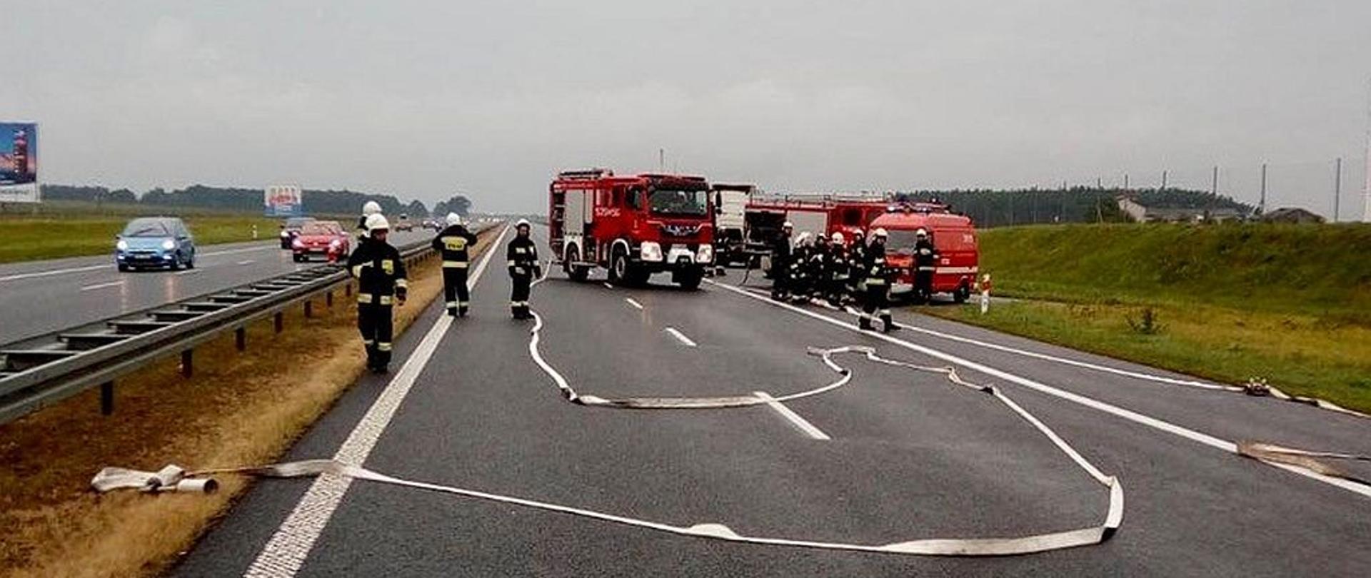 Zdjęcie przedstawia ratowników na autostradzie, w tle widać pojazdy uczestniczące w wypadku, rozwinięte węże, kilka pojazdów straży pożarnej