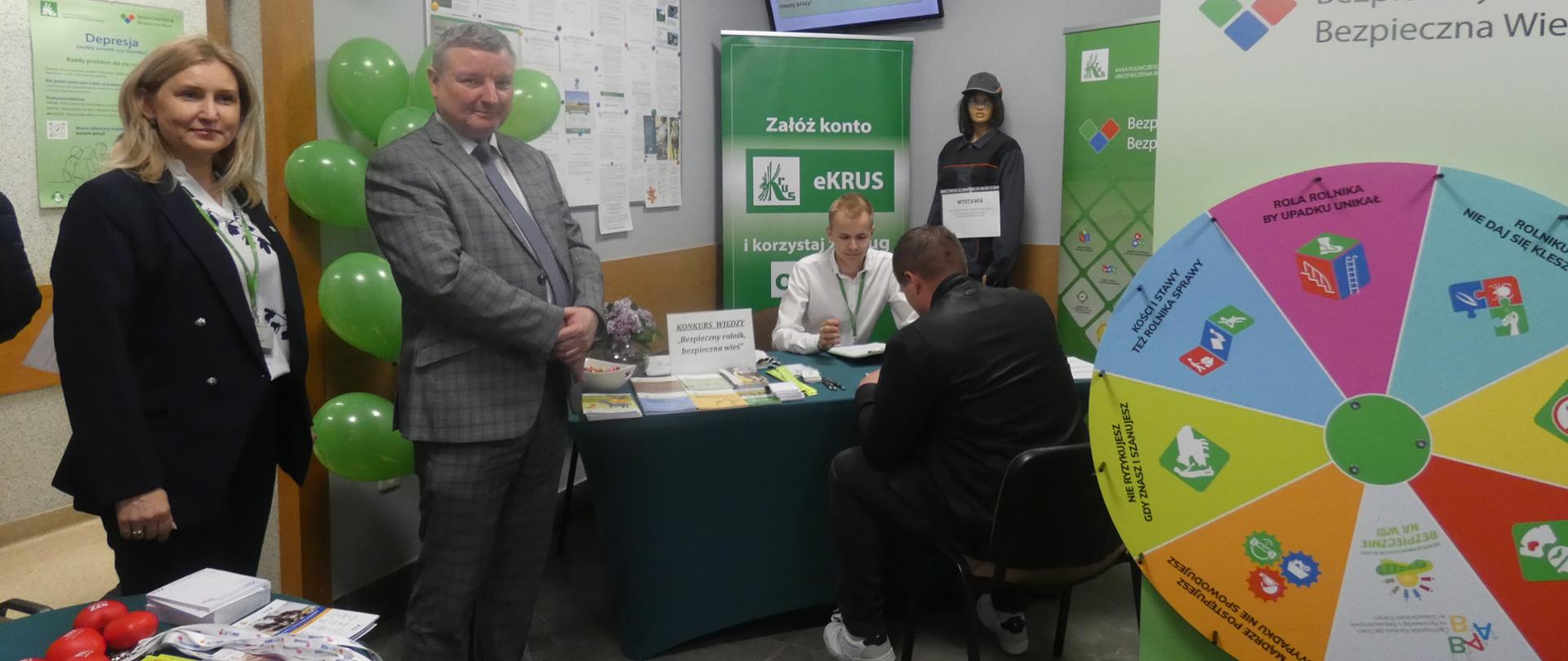 Zdjęcie przedstawia cztery osoby na sali obsługi interesanta w Placówce Terenowej KRUS w Wysokiem Mazowieckiem – dwie stojące po lewej stronie (kobieta i mężczyzna w stroju służbowym), mężczyzna siedzący przy stoliku oraz kolejny mężczyzna naprzeciwko, wypełniający formularz konkursowy. W tle widoczny kolorowy baner promujący eKRUS, stanowisko konkursowe z materiałami informacyjnymi, balony w kolorze zielonym oraz kolorowe Koło Bezpieczeństwa z hasłami dotyczącymi bezpiecznej pracy w rolnictwie.