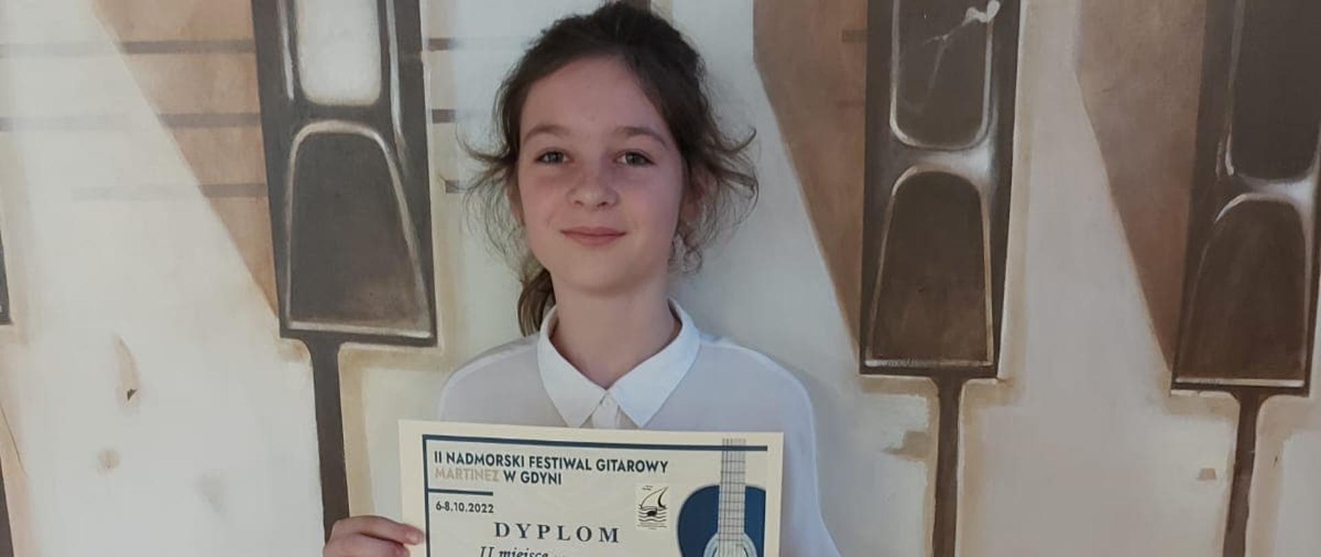Uczennica Urszula Wachowska prezentuje dyplom za zdobycie drugiego miejsca w Nadmorskim Festiwalu Gitarowym MARTINEI w Gdyni na tle artystycznego obrazu klawiatury fortepianowej i nut.