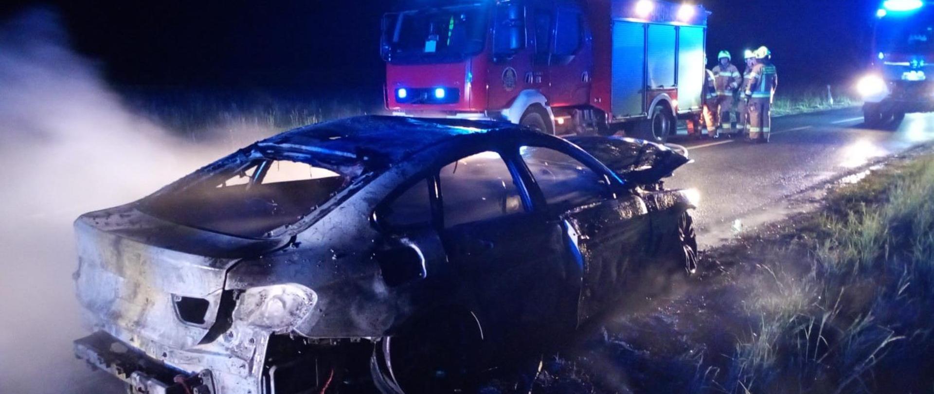 Na zdjęciu widać całkowicie spalony samochód osobowy, który stoi na poboczu drogi. Brakuje tylnego zderzaka oraz reflektorów, tylna część nadwozia jest mocno zdeformowana. W tle widoczne są dwa wozy strażackie z włączonymi niebieskimi światłami ostrzegawczymi. Pięciu strażaków w pełnym umundurowaniu znajduje się obok jednego z pojazdów ratowniczych. Jest ciemno, ale miejsce akcji jest oświetlone.