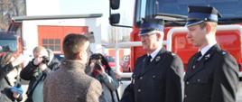 Wójt gminy Kunów wręcza akt przekazania pojazdu prezesowi OSP Bałtów. W tle pozyskany samochód gaśniczy i fotoreporterzy wykonujący pamiątkowe zdjęcia.